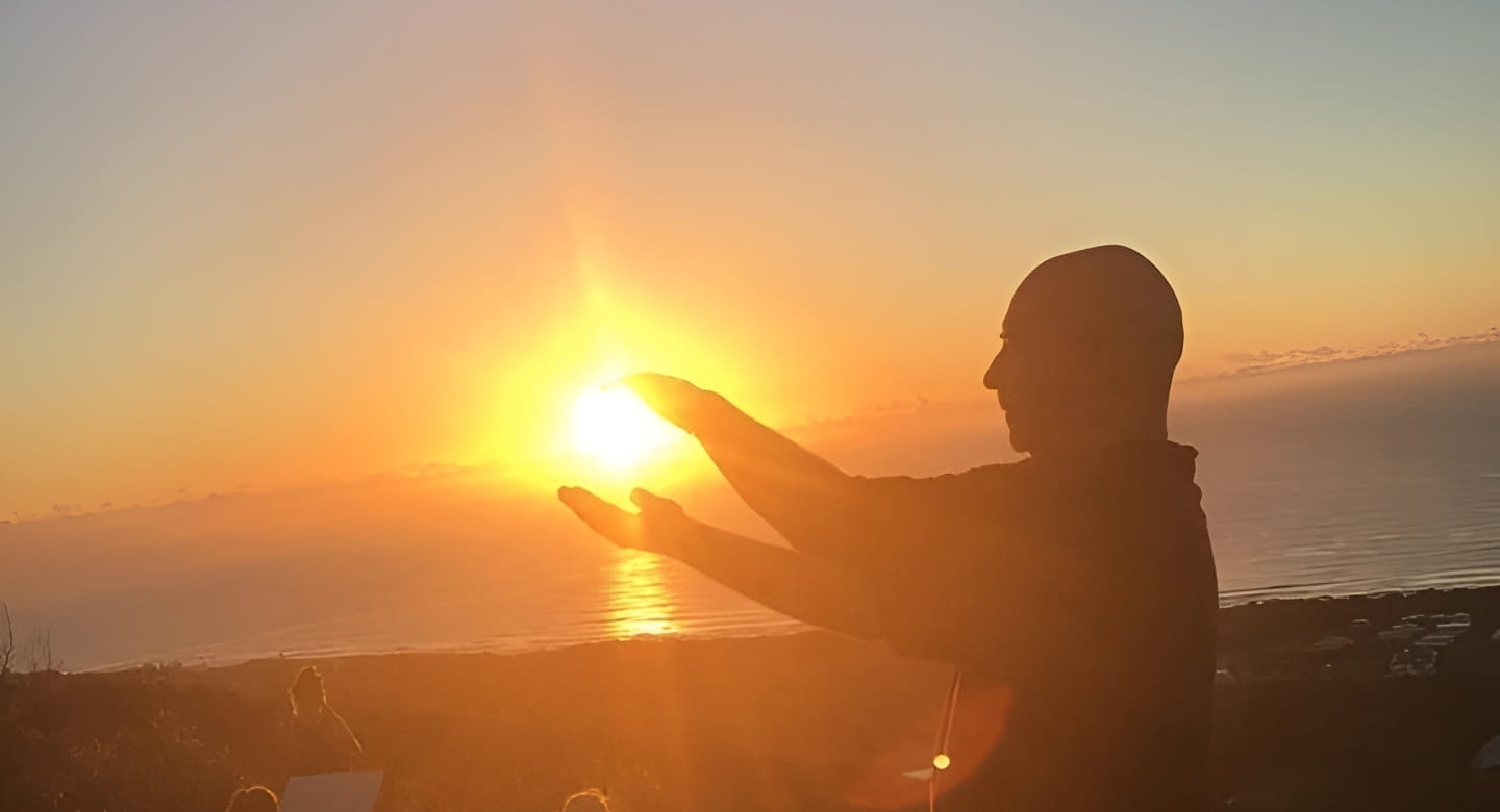 Kadama in silhouette at sunrise, arms extended in a qigong posture above a small group of practitioners on a clifftop overlooking the ocean